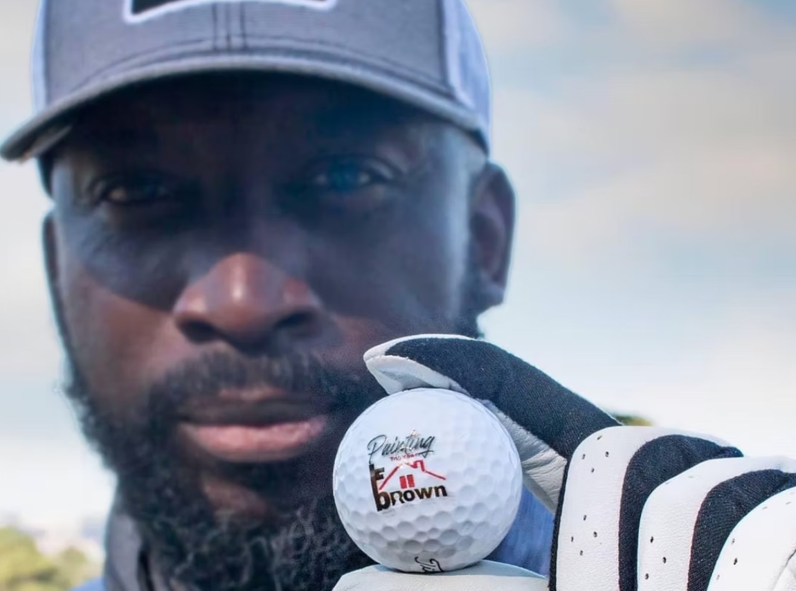 Photograph of a person holding a white golf ball featuring Painting the Town Brown logo
